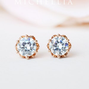 May include: A pair of rose gold stud earrings with a clear round stone set in a delicate, ornate, gold setting.  MICHELLIA is written in the background.