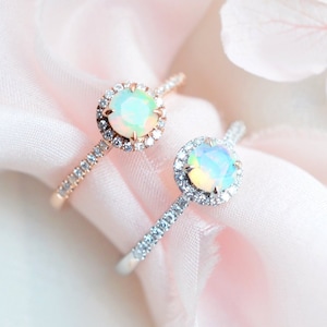 May include: Two rings with opal gemstones surrounded by a halo of smaller diamonds. The ring on the left has a rose gold band, while the ring on the right has a silver band. Both rings are on a pink fabric background.