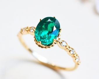 Op de afbeelding: Een gouden ring met een grote ovale smaragdgroene edelsteen, omringd door kleinere heldere edelstenen. De ring ligt op een witte achtergrond.