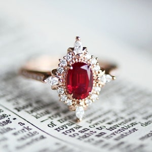 Pode incluir: Um anel de noivado de rubi vermelho com um halo de diamantes engastado em uma banda de ouro rosa. O anel está sobre um livro com a palavra "royal" visível.