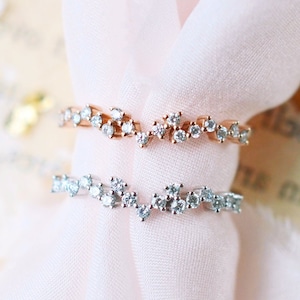 Pu&ograve; includere: Due anelli delicati con un design a forma di onda incastonati con piccoli diamanti scintillanti. Gli anelli sono su uno sfondo di tessuto rosa morbido.