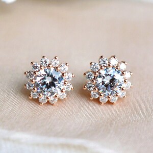 May include: A pair of rose gold stud earrings with a round, clear crystal center surrounded by a halo of smaller crystals.