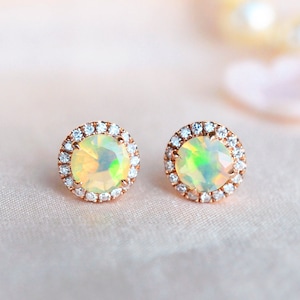 May include: A pair of rose gold earrings with a round opal gemstone surrounded by a halo of small clear cubic zirconia stones.