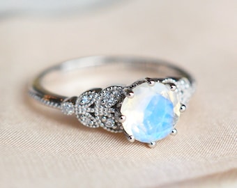 Op de afbeelding: Een zilveren ring met een grote, ronde, iriserende edelsteen omringd door kleinere, heldere edelstenen. De ring heeft een delicaat, bloemig ontwerp.