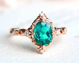 Op de afbeelding: Een ring van roségoud met een grote ovale smaragdgroene edelsteen, omringd door kleinere heldere edelstenen. De ring heeft een delicaat, ingewikkeld ontwerp.