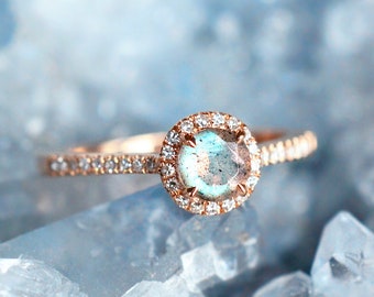 Op de afbeelding: Een ring van roségoud met een ronde, iriserende blauwe edelsteen omringd door een halo van kleinere diamanten. De ring ligt op een bed van witte kristallen.