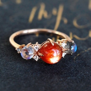 May include: A rose gold ring with three stones: a large sunstone, a smaller moonstone, and a clear stone. The ring is set on a dark background.