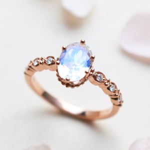 May include: A rose gold ring with a large oval moonstone and smaller clear stones set in a delicate band. The ring is on a white background with pink flower petals.