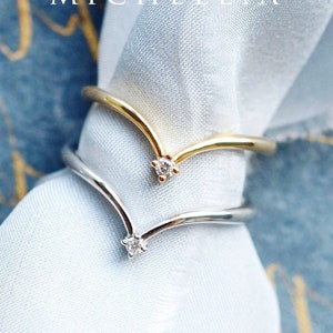 May include: Two gold and silver V-shaped rings with a small diamond at the point of the V. The rings are on a light blue fabric background.