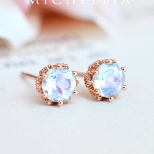 May include: A pair of rose gold earrings with a round, iridescent gemstone set in a delicate, ornate frame. The earrings are on a white background.