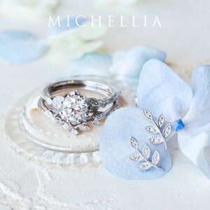 Op de afbeelding: Een delicate zilveren ring met een sprankelende diamant omringd door een bloemmotief. De ring wordt getoond op een wit oppervlak met blauwe bloemblaadjes. De tekst "MICHELLIA" is zichtbaar op de achtergrond.