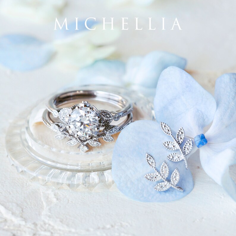 Op de afbeelding: Een delicate zilveren ring met een sprankelende diamant omringd door een bloemmotief. De ring wordt getoond op een wit oppervlak met blauwe bloemblaadjes. De tekst "MICHELLIA" is zichtbaar op de achtergrond.