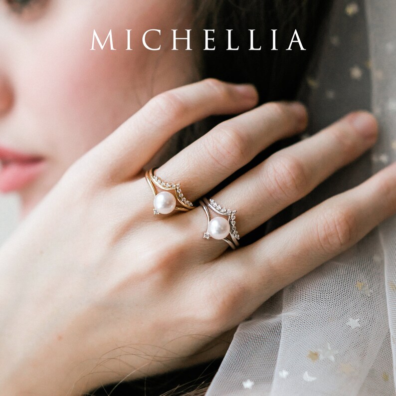 May include: Two gold and silver rings with a pearl on each. The rings are stacked on a finger and have a delicate, v-shaped design with small diamonds.