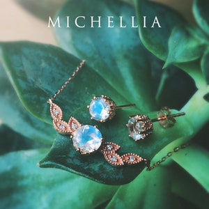 May include: A delicate rose gold necklace and earrings set with a moonstone pendant and matching earrings. The jewelry is set against a green leafy background.