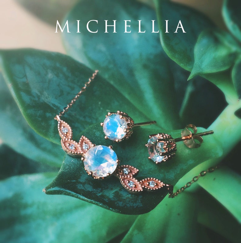 May include: A delicate rose gold necklace and earrings set with a moonstone pendant and matching earrings. The jewelry is set against a green leafy background.