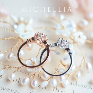 May include: Two delicate rings with floral designs, one in rose gold and one in silver, are displayed on a background of white pearls and a piece of paper with the word "MICHELLIA" printed on it.