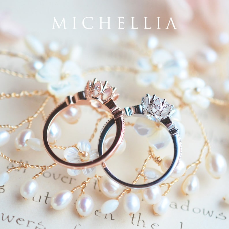 May include: Two delicate rings with floral designs, one in rose gold and one in silver, are displayed on a background of white pearls and a piece of paper with the word "MICHELLIA" printed on it.