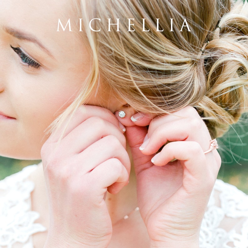 May include: A woman is putting on a small, round, sparkly earring. The earring is silver and has a small, clear stone in the center. The woman is wearing a white dress and has her hair pulled back in a bun.