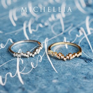 Pu&ograve; includere: Due anelli delicati con un designo di diamanti scintillanti. Gli anelli sono su uno sfondo blu con scrittura bianca in corsivo. La parola "MICHELLIA" &egrave; scritta in bianco nella parte superiore dell'immagine.