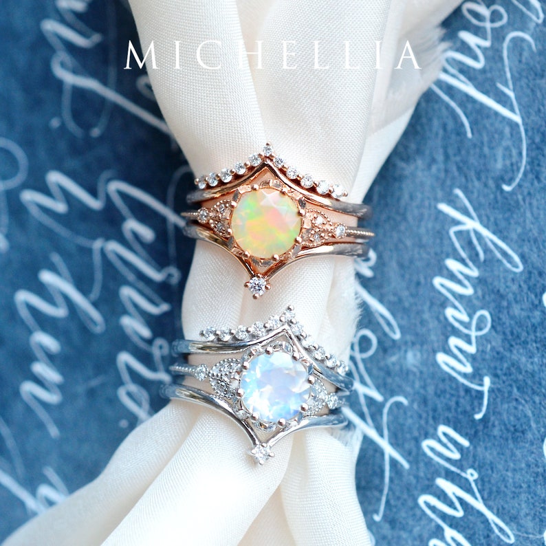 May include: Two delicate rings with opal gemstones set in a pointed crown design. The top ring is rose gold with a pale yellow opal, and the bottom ring is silver with a white opal.