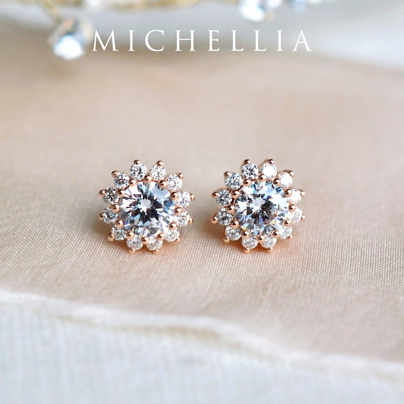May include: A pair of rose gold stud earrings with a round, clear crystal center surrounded by a halo of smaller crystals.
