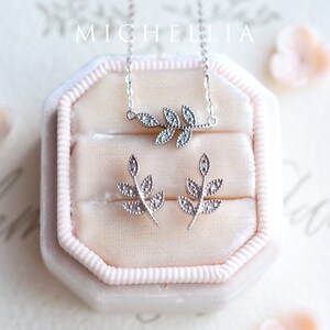Op de afbeelding: Een zilveren ketting- en oorstekerset met een delicaat takontwerp met glinsterende zirkonia-stenen. De sieraden worden gepresenteerd in een roze fluwelen doos.