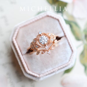 May include: A rose gold engagement ring set with a round brilliant cut diamond surrounded by smaller diamonds. The ring features a delicate floral design with leaves and vines.