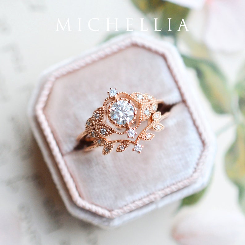 May include: A rose gold engagement ring set with a round brilliant cut diamond surrounded by smaller diamonds. The ring features a delicate floral design with leaves and vines.