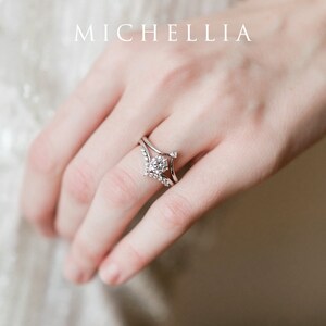 May include: A silver ring with a large round diamond center stone and smaller diamonds set in a V-shaped band. The ring is worn on the ring finger of a hand.