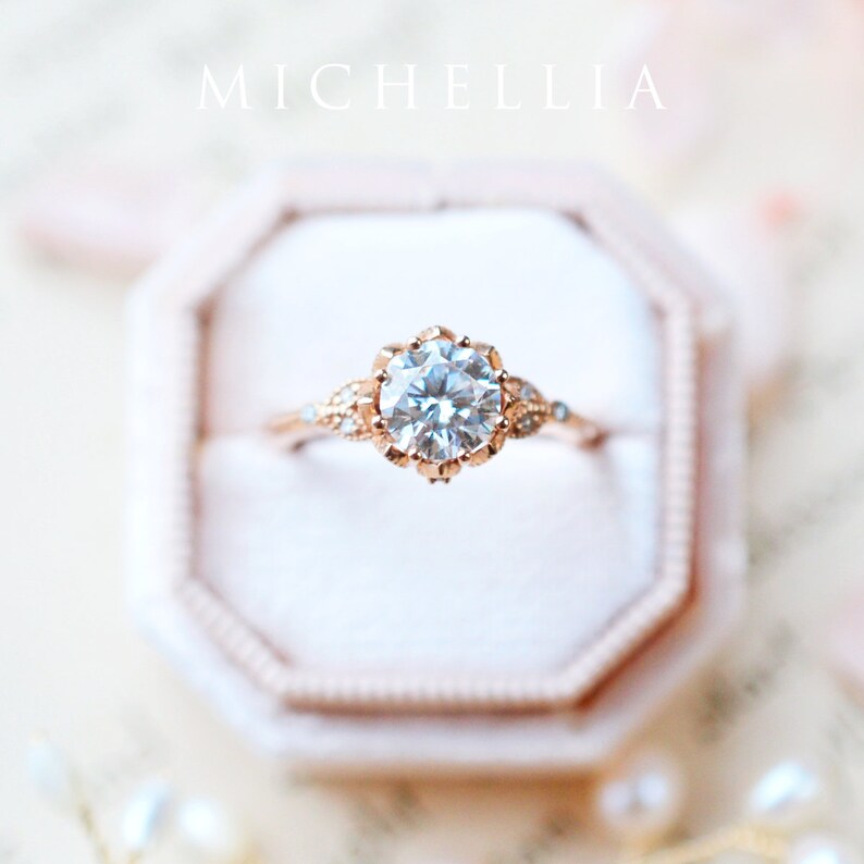 May include: A delicate rose gold engagement ring with a round brilliant cut diamond centre stone. The ring features a floral design with small diamonds on the band.