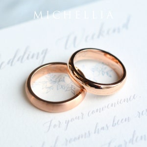 May include: Two rose gold wedding bands are displayed on a white surface with elegant script. The rings are smooth and shiny, with a classic, rounded design. The word "MICHELLIA" is at the top of the image.