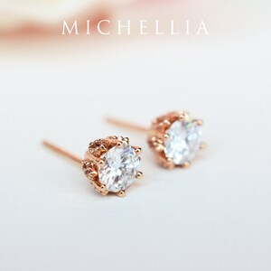 May include: A pair of rose gold stud earrings with a clear round stone set in a decorative crown-like setting.