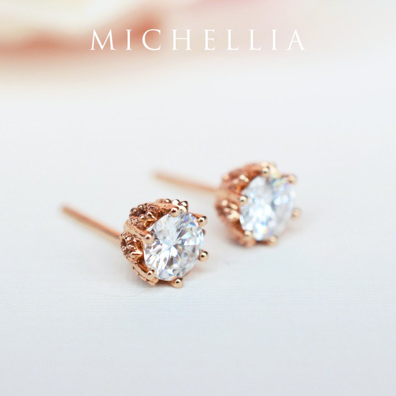 May include: A pair of rose gold stud earrings with a clear round stone set in a decorative crown-like setting.