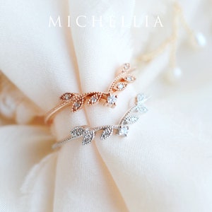 May include: Two delicate rings with a leaf design and small clear stones. The rings are on a white fabric background.