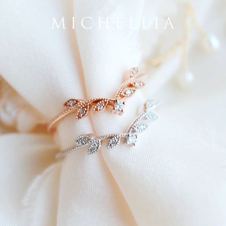 May include: Two delicate rings with a leaf design and small clear stones. The rings are on a white fabric background.