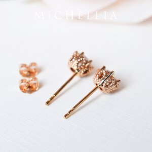 May include: A pair of rose gold stud earrings with a small, clear stone set in a delicate, textured crown-like setting. The earrings have a post back.