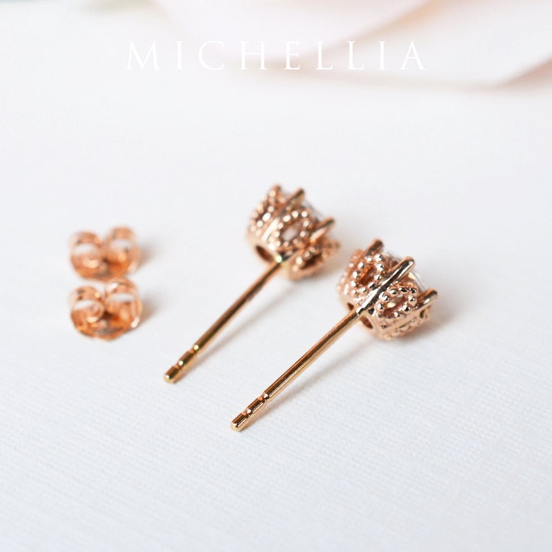 May include: A pair of rose gold stud earrings with a small, clear stone set in a delicate, textured crown-like setting. The earrings have a post back.