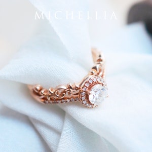 K&ouml;nnte beinhalten: Ein Verlobungsring aus Ros&eacute;gold mit einem gro&szlig;en, runden, klaren Diamanten, der von kleineren Diamanten umgeben ist. Der Ring hat ein zartes, verziertes Design.
