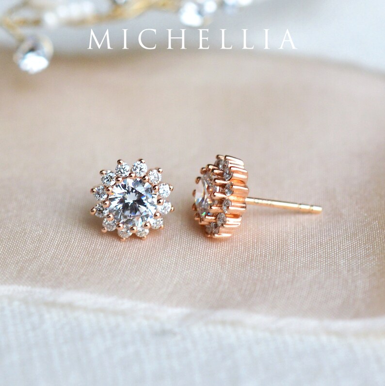 May include: A pair of rose gold stud earrings with a round, clear crystal center surrounded by smaller crystals. The earrings have a simple, elegant design.