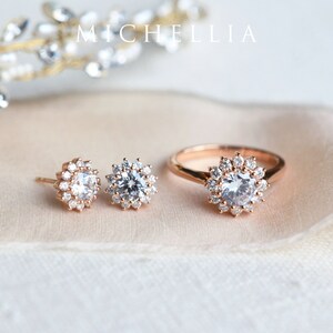 May include: A set of rose gold jewelry featuring a ring and a pair of earrings. Each piece has a round, clear stone surrounded by a halo of smaller stones.