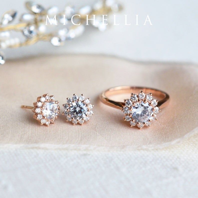 May include: A set of rose gold jewelry featuring a ring and a pair of earrings. Each piece has a round, clear stone surrounded by a halo of smaller stones.