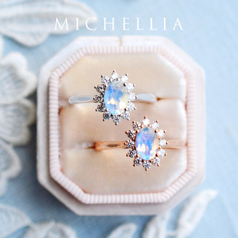 May include: Two oval-shaped moonstone rings with diamond halo settings. The ring on the left is set in silver, and the ring on the right is set in rose gold. Both rings are in a white velvet ring box.