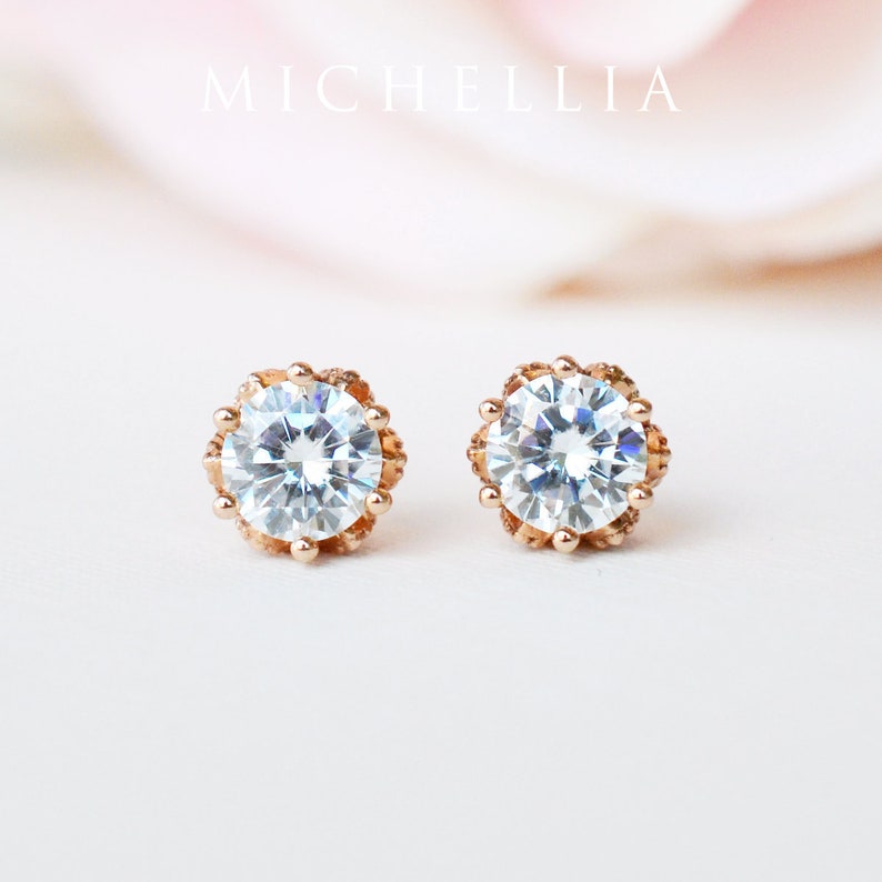 May include: A pair of rose gold stud earrings with a clear round stone set in a delicate, ornate, gold setting.  MICHELLIA is written in the background.