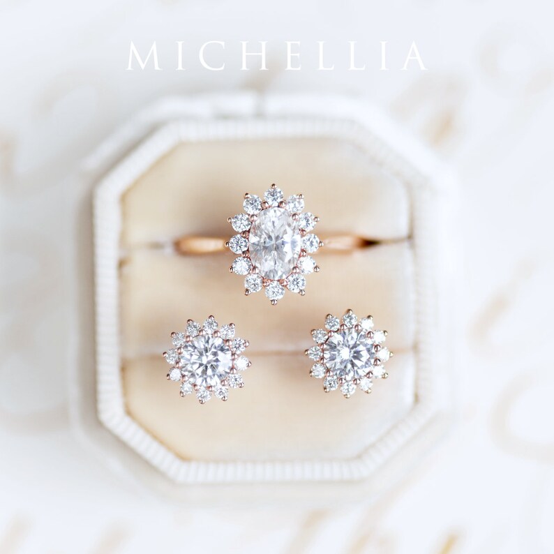 May include: A set of three rose gold rings with a large oval-shaped diamond in the center of the engagement ring and smaller round diamonds in the center of the wedding bands. The rings are displayed in a white ring box.