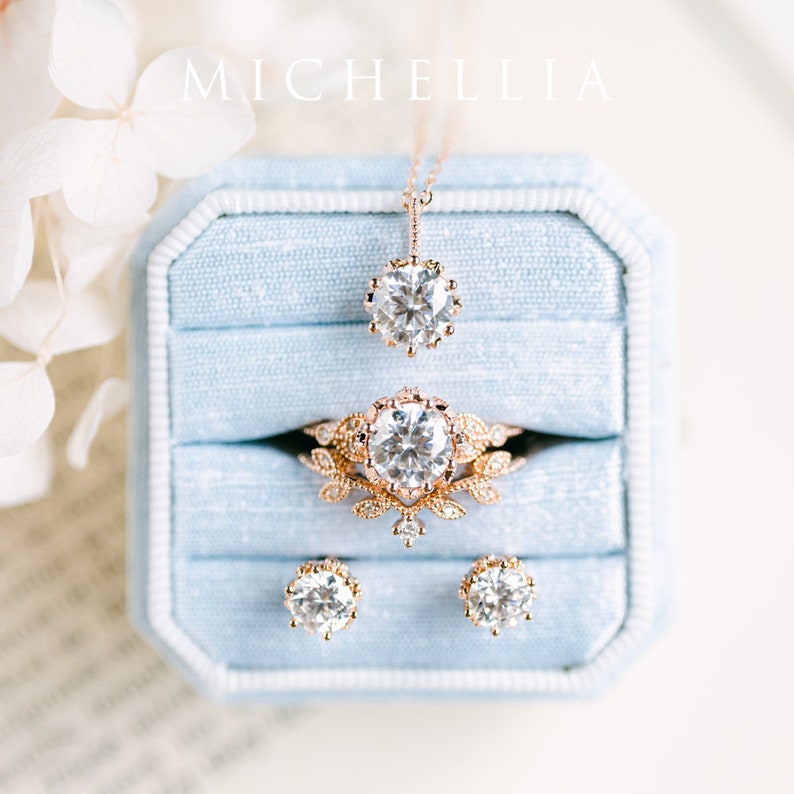 May include: A set of delicate gold jewellery with a floral design, including a necklace, ring, and earrings. The jewellery is displayed in a light blue velvet box.