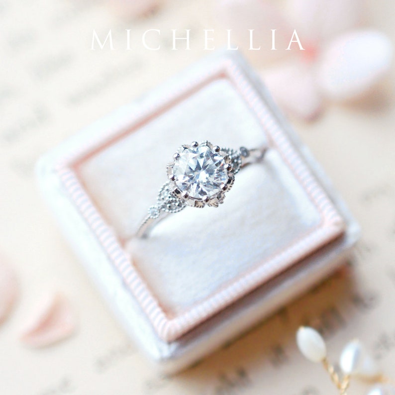 May include: A silver engagement ring featuring a large, clear, round diamond set in a delicate, ornate setting. The ring is displayed in a white velvet ring box.