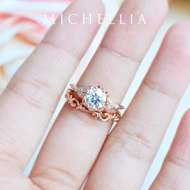 K&ouml;nnte beinhalten: Ein Verlobungsring aus Ros&eacute;gold mit einem gro&szlig;en runden Mittelst&uuml;ck und kleineren Akzent-Steinen. Der Ring ist auf einem zarten Band mit einem floralen Design gesetzt.