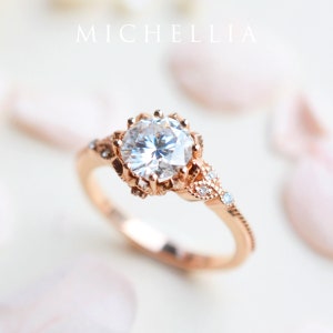 May include: A rose gold engagement ring featuring a large, clear, round-cut centre stone surrounded by smaller stones. The band is adorned with delicate floral details.