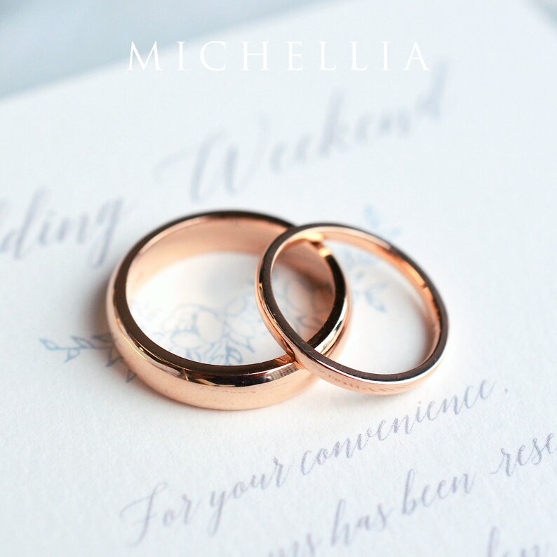 May include: Two rose gold wedding rings, one wider than the other, rest on a white surface with faint script and floral designs. The word "MICHELLIA" is at the top. The rings are the focal point, symbolizing commitment and love.