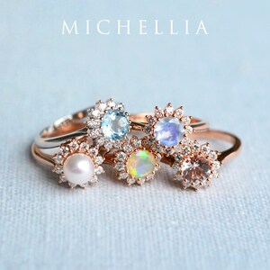May include: Five rings with different gemstones set in a halo of diamonds. The rings are stacked on top of each other. The gemstones are aquamarine, moonstone, opal, morganite, and pearl.